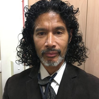 Dr Andrew Britto smiles at the camera. He is wearing a black suit and has a stethoscope around his neck